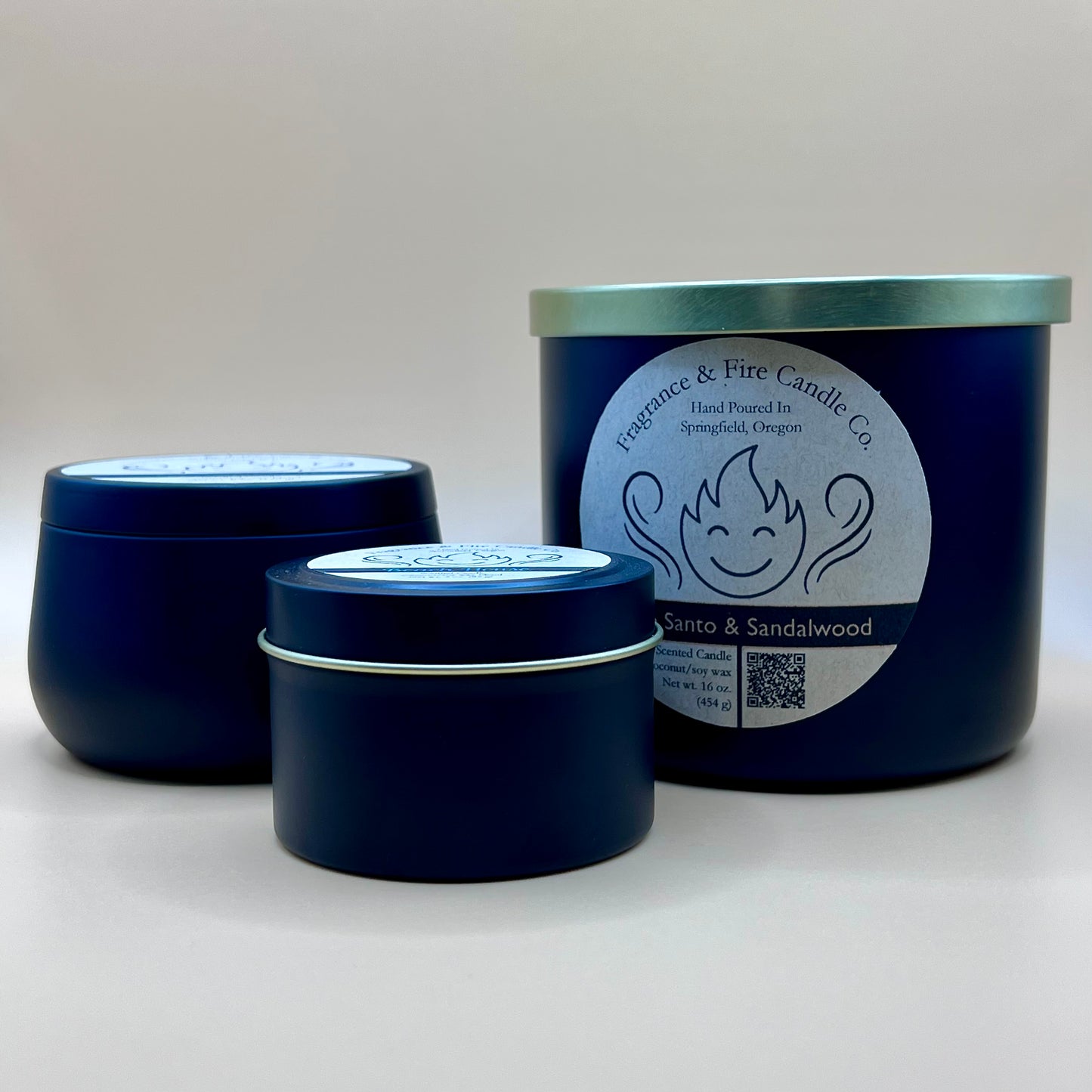 Three black candles from Fragrance & Fire Candle Co., Springfield, Oregon, in different sizes. The largest candle on the right is a 16oz glass container with a gold lid and a white label featuring a stylized flame logo, the text "Fragrance & Fire Candle Co. Hand Poured In Springfield, Oregon," and "Palo Santo & Sandalwood" scent name. Details include "Scented Candle Coconut/soy wax Net wt. 16 oz (454 g)" and a QR code. The two smaller candles on the left are black tins with light-colored labels.