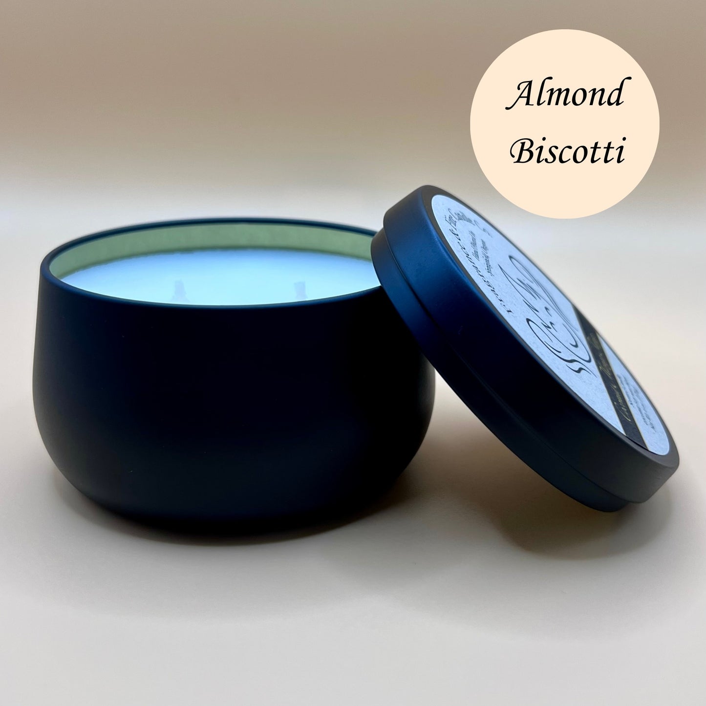 6oz black tin candle from Fragrance & Fire Candle Co., Springfield, Oregon, with an open lid resting beside it. The tin contains white wax. An almond colored circle in the upper right corner reads "Almond Biscotti." The open lid shows a partial label design.