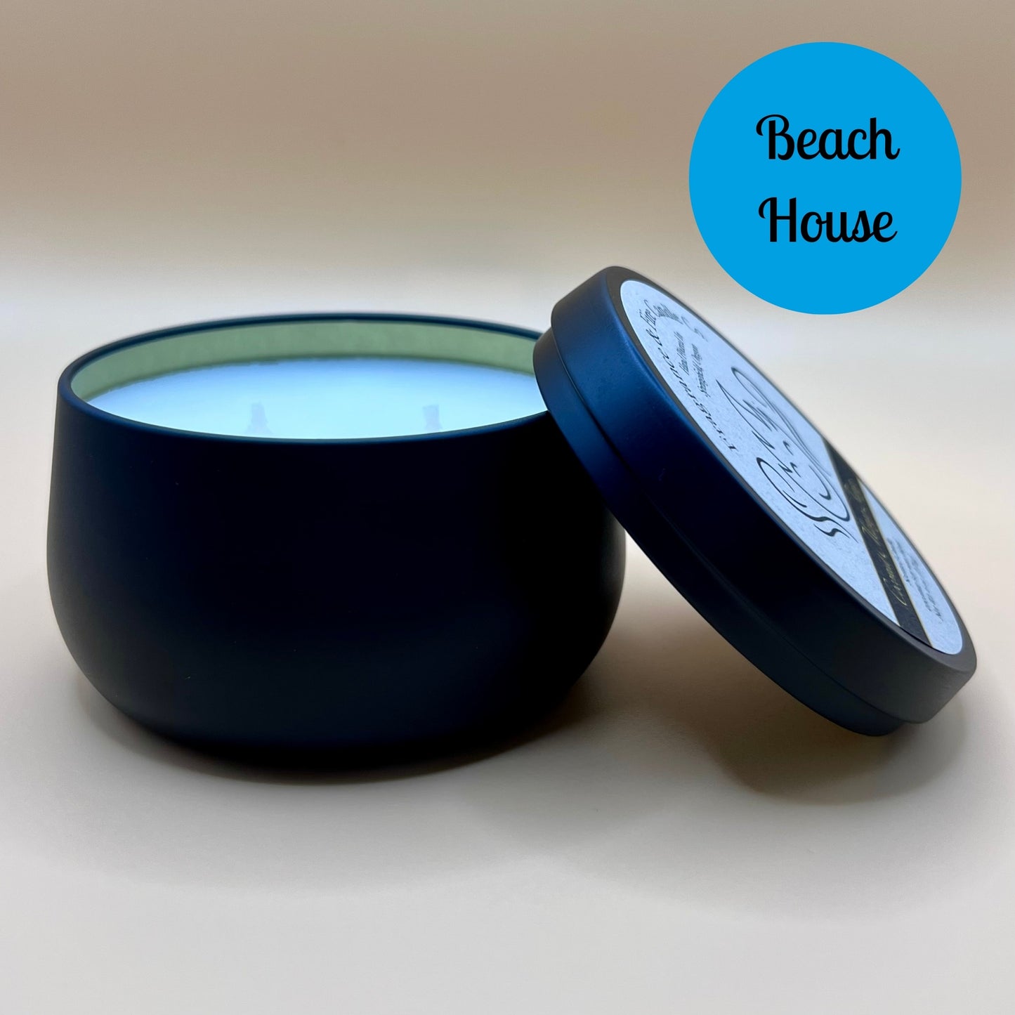 6oz black tin candle from Fragrance & Fire Candle Co., Springfield, Oregon, with an open lid resting beside it. The tin contains white wax. A blue circle in the upper right corner reads "Beach House." The open lid shows a partial label design.