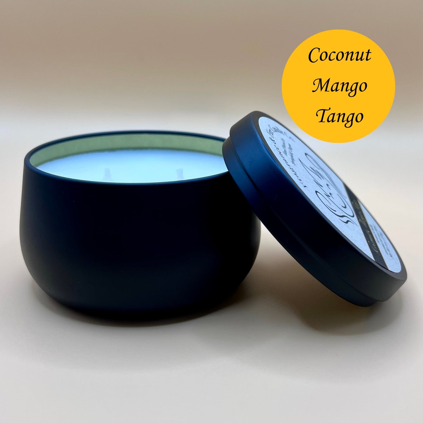 6oz black tin candle from Fragrance & Fire Candle Co., Springfield, Oregon, with an open lid resting beside it. The tin contains white wax. A yellow circle in the upper right corner reads "Coconut Mango Tango." The open lid shows a partial label design.