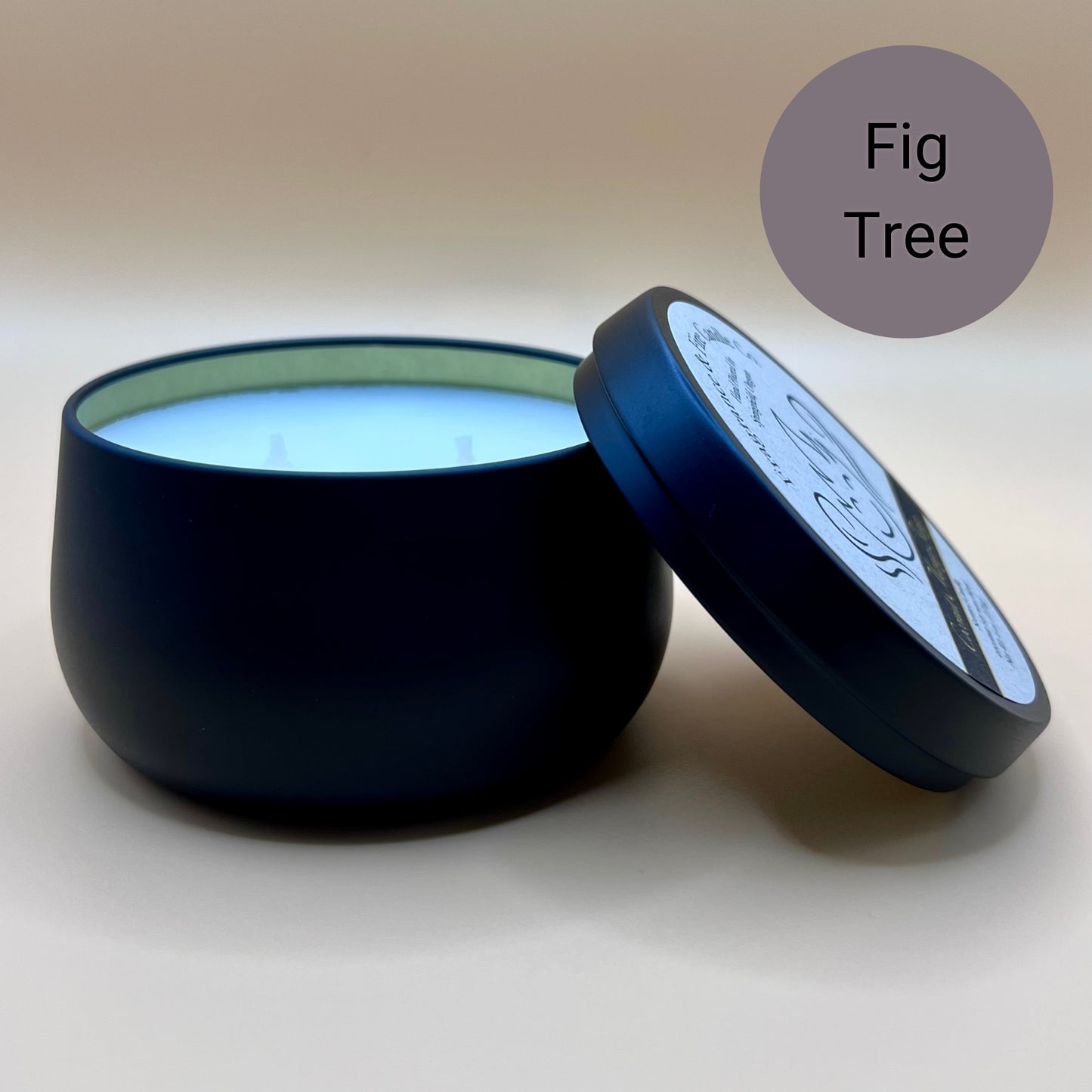 6oz black tin candle from Fragrance & Fire Candle Co., Springfield, Oregon, with an open lid resting beside it. The tin contains white wax. A grayish purple circle in the upper right corner reads "Fig Tree." The open lid shows a partial label design.