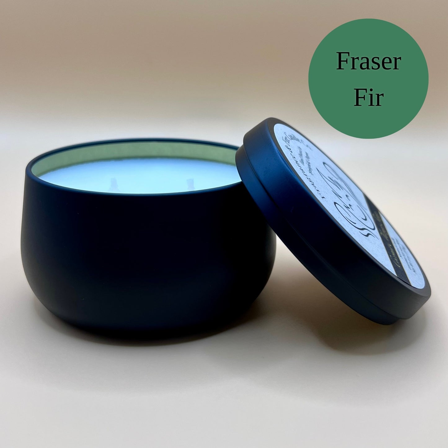 6oz black tin candle from Fragrance & Fire Candle Co., Springfield, Oregon, with an open lid resting beside it. The tin contains white wax. A green circle in the upper right corner reads "Fraser Fir." The open lid shows a partial label design.