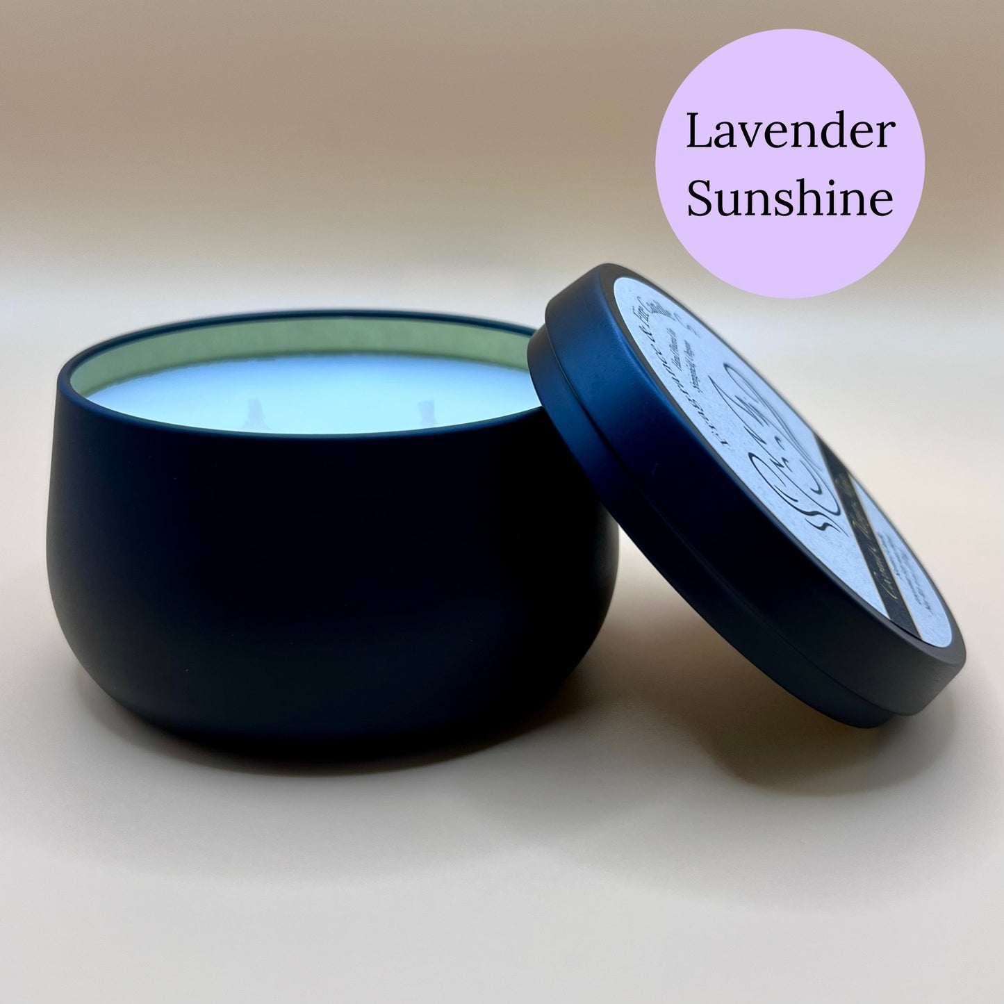 6oz black tin candle from Fragrance & Fire Candle Co., Springfield, Oregon, with an open lid resting beside it. The tin contains white wax. A light purple circle in the upper right corner reads "Lavender Sunshine." The open lid shows a partial label design.