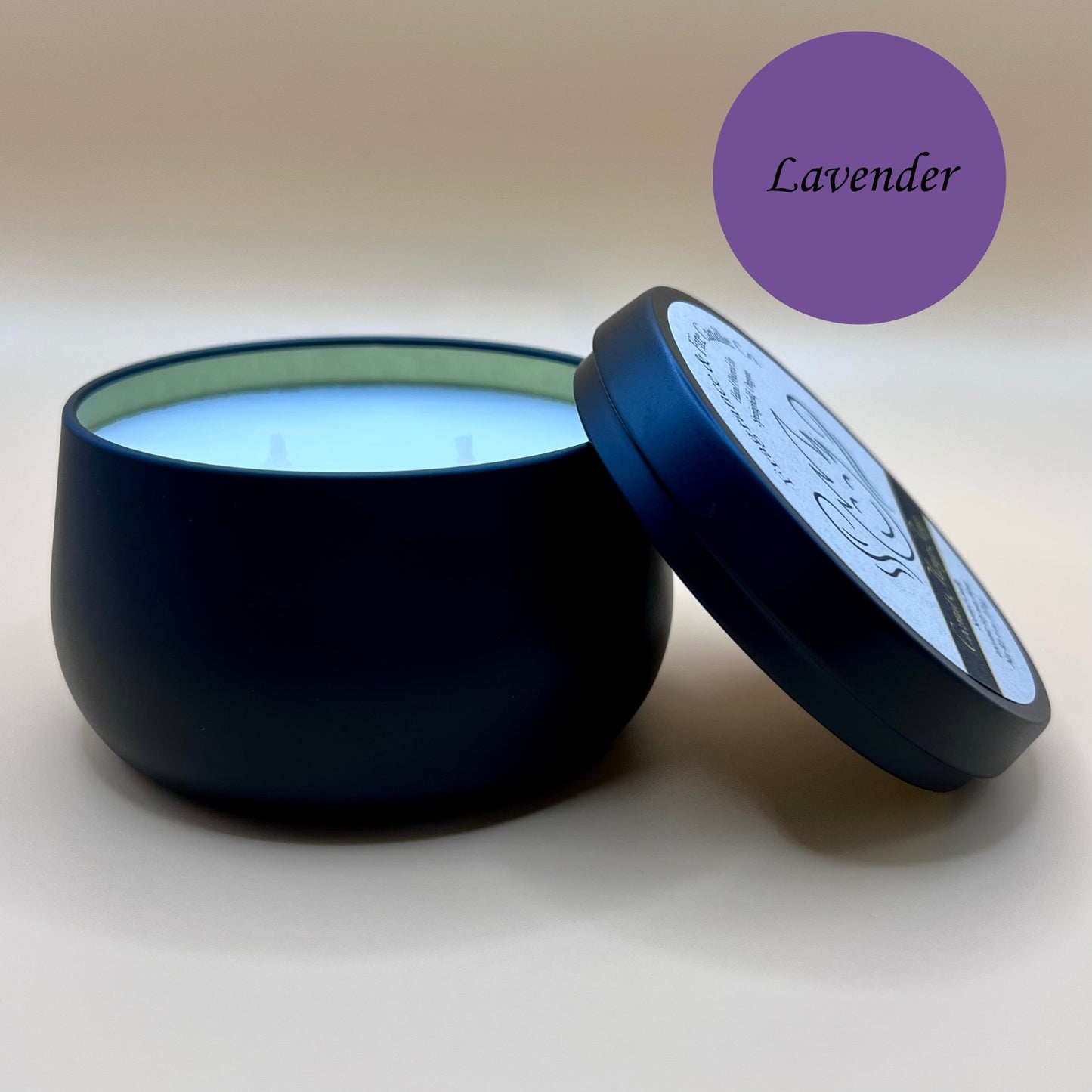 6oz black tin candle from Fragrance & Fire Candle Co., Springfield, Oregon, with an open lid resting beside it. The tin contains white wax. A purple circle in the upper right corner reads "Lavender" The open lid shows a partial label design.
