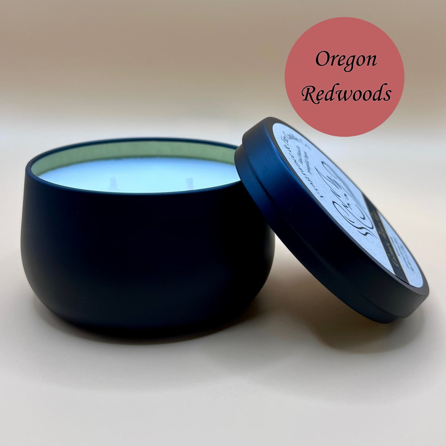 6oz black tin candle from Fragrance & Fire Candle Co., Springfield, Oregon, with an open lid resting beside it. The tin contains white wax. A burnt red circle in the upper right corner reads "Oregon Redwoods." The open lid shows a partial label design.