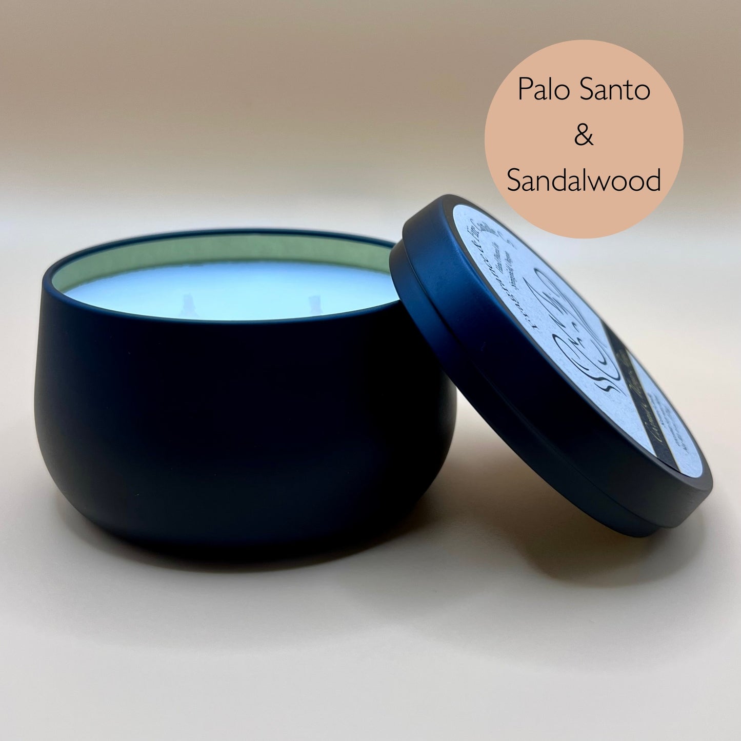 6oz black tin candle from Fragrance & Fire Candle Co., Springfield, Oregon, with an open lid resting beside it. The tin contains white wax. A light brown circle in the upper right corner reads "Palo Santo & Sandalwood." The open lid shows a partial label design.