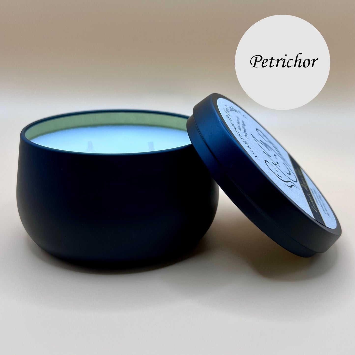 6oz black tin candle from Fragrance & Fire Candle Co., Springfield, Oregon, with an open lid resting beside it. The tin contains white wax. A light circle in the upper right corner reads "Petrichor." The open lid shows a partial label design.