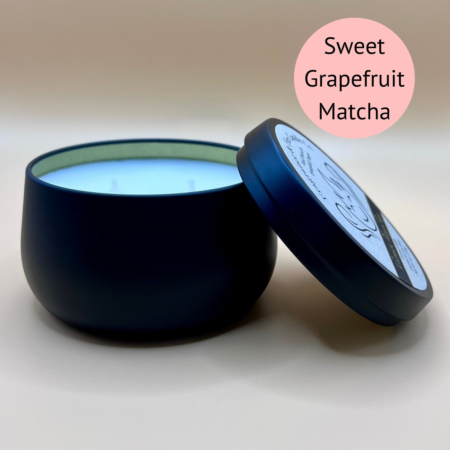 6oz black tin candle from Fragrance & Fire Candle Co., Springfield, Oregon, with an open lid resting beside it. The tin contains white wax. A light pink circle in the upper right corner reads "Sweet Grapefruit Matcha." The open lid shows a partial label design.