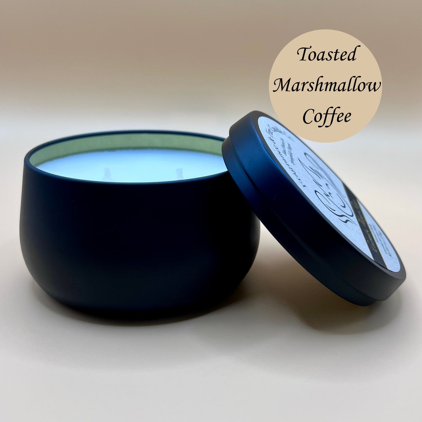 6oz black tin candle from Fragrance & Fire Candle Co., Springfield, Oregon, with an open lid resting beside it. The tin contains white wax. A tan circle in the upper right corner reads "Toasted Marshmallow Coffee." The open lid shows a partial label design.