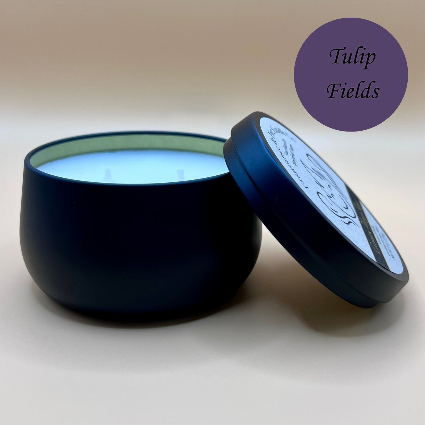 6oz black tin candle from Fragrance & Fire Candle Co., Springfield, Oregon, with an open lid resting beside it. The tin contains white wax. A purple circle in the upper right corner reads "Tulip Fields." The open lid shows a partial label design.