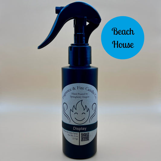 Black spray bottle from Fragrance & Fire Candle Co., Springfield, Oregon, with a black spray nozzle. The bottle has a white label with a stylized flame logo, the text "Fragrance & Fire Candle Co. Hand Poured In Springfield, Oregon," and "Display" scent name. The label also includes a QR code and the text "Room & Linen Spray Net Wt. 4 oz (113g)." A blue circle in the upper right corner reads "Beach House."
