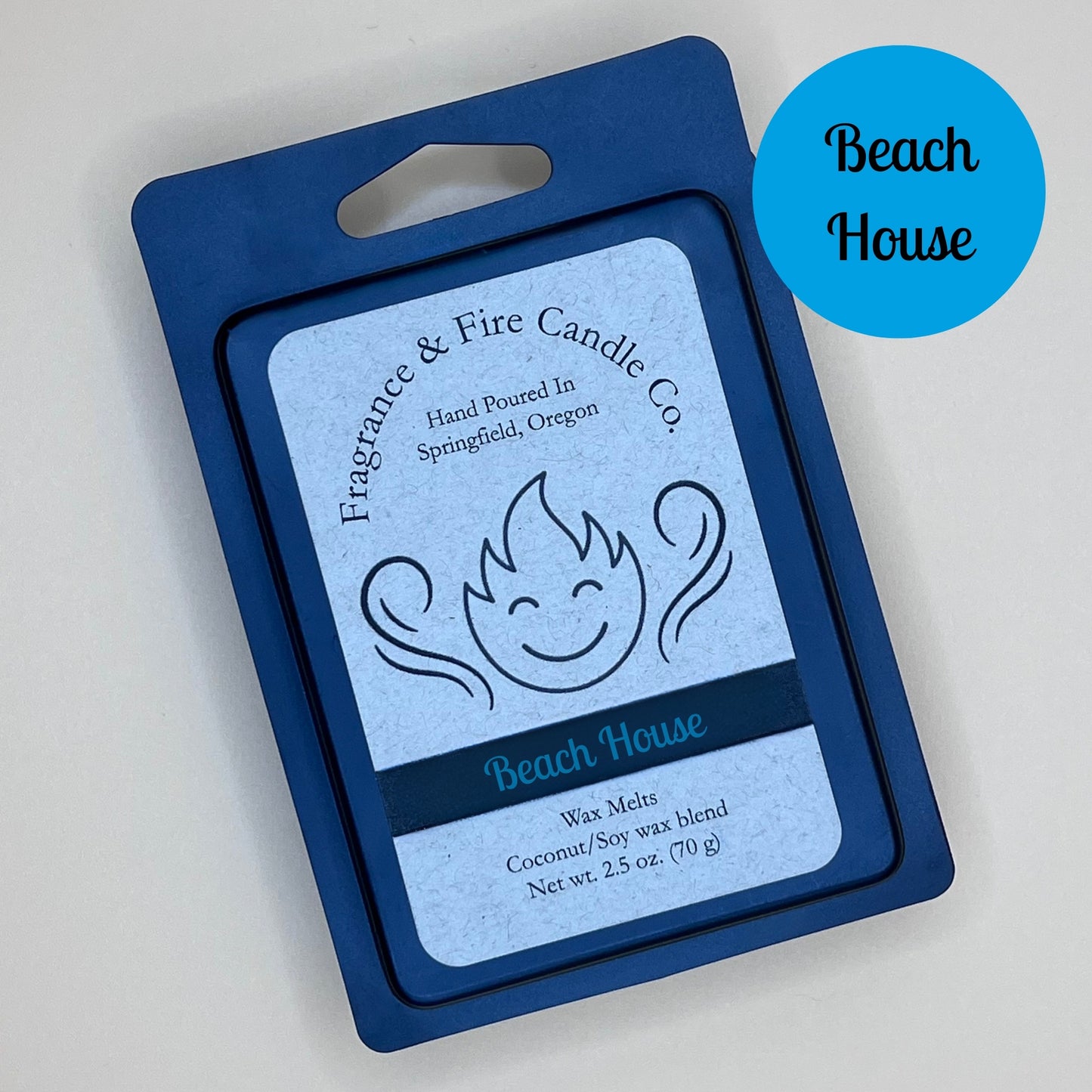 Black clamshell packaging for wax melts from Fragrance & Fire Candle Co., Springfield, Oregon. The packaging features a white label with a stylized flame logo, the text "Fragrance & Fire Candle Co. Hand Poured In Springfield, Oregon," and "Beach house" scent name. The label also includes the text "Wax Melts Coconut/Soy wax blend Net wt. 2.5 oz. (70 g)." A blue circle in the upper right corner says scent name.