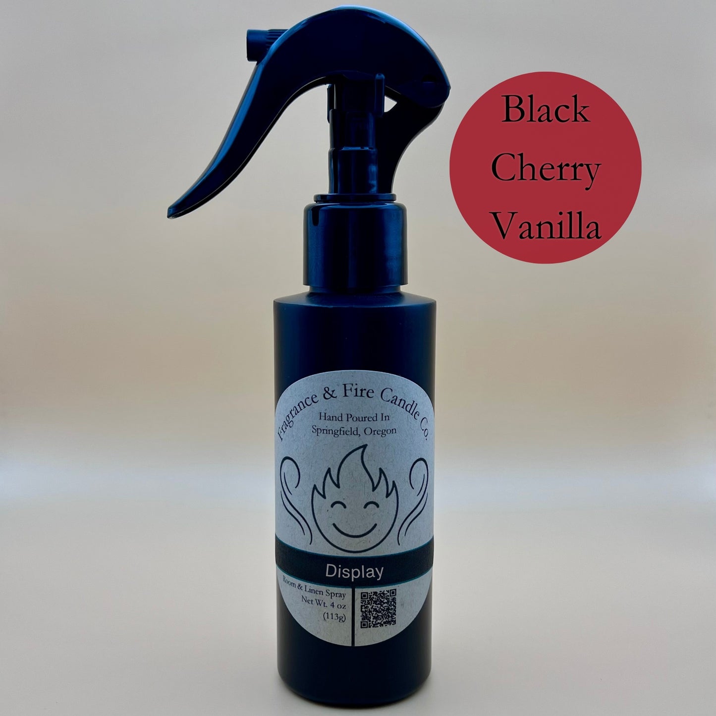 Black spray bottle from Fragrance & Fire Candle Co., Springfield, Oregon, with a black spray nozzle. The bottle has a white label with a stylized flame logo, the text "Fragrance & Fire Candle Co. Hand Poured In Springfield, Oregon," and "Display" scent name. The label also includes a QR code and the text "Room & Linen Spray Net Wt. 4 oz (113g)." A dark red circle in the upper right corner reads "Black Cherry Vanilla."