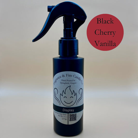 Black spray bottle from Fragrance & Fire Candle Co., Springfield, Oregon, with a black spray nozzle. The bottle has a white label with a stylized flame logo, the text "Fragrance & Fire Candle Co. Hand Poured In Springfield, Oregon," and "Display" scent name. The label also includes a QR code and the text "Room & Linen Spray Net Wt. 4 oz (113g)." A dark red circle in the upper right corner reads "Black Cherry Vanilla."