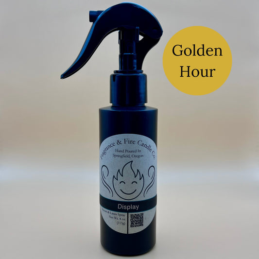 Black spray bottle from Fragrance & Fire Candle Co., Springfield, Oregon, with a black spray nozzle. The bottle has a white label with a stylized flame logo, the text "Fragrance & Fire Candle Co. Hand Poured In Springfield, Oregon," and "Display" scent name. The label also includes a QR code and the text "Room & Linen Spray Net Wt. 4 oz (113g)." A dark yellow circle in the upper right corner reads "Golden Hour."