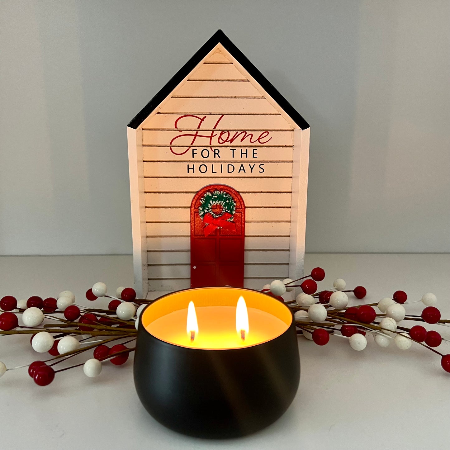 Lit Fragrance & Fire candle in front of christmas berries decor and a house shaped sign that reads "Home for the holidays".