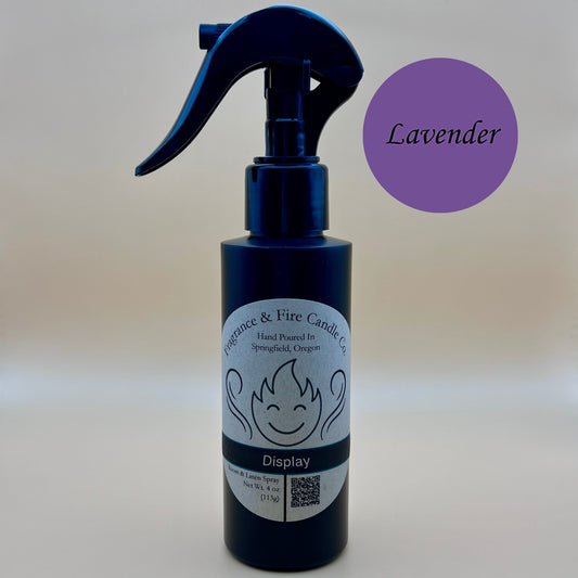 Black spray bottle from Fragrance & Fire Candle Co., Springfield, Oregon, with a black spray nozzle. The bottle has a white label with a stylized flame logo, the text "Fragrance & Fire Candle Co. Hand Poured In Springfield, Oregon," and "Display" scent name. The label also includes a QR code and the text "Room & Linen Spray Net Wt. 4 oz (113g)." A purple circle in the upper right corner reads "Lavender."