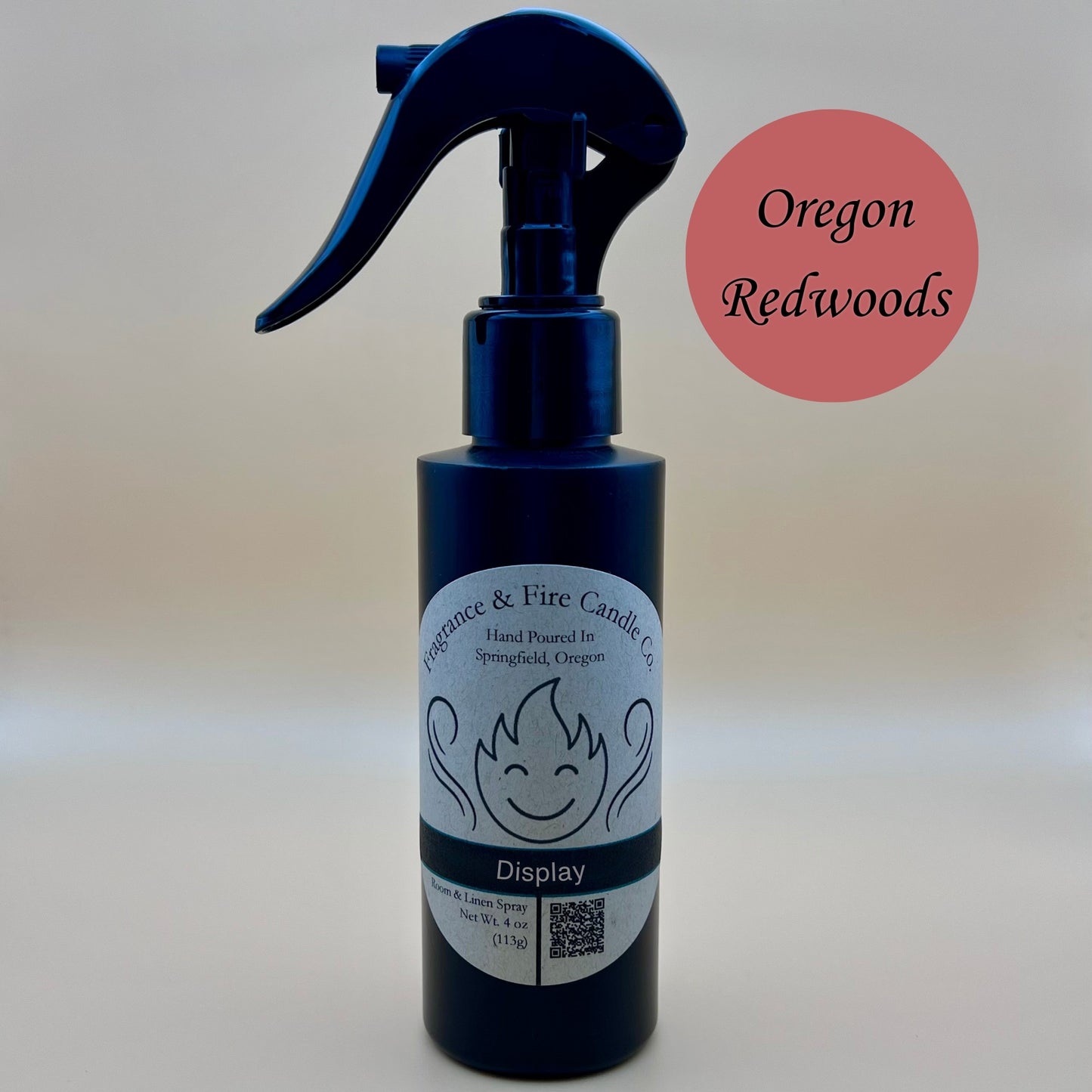 Black spray bottle from Fragrance & Fire Candle Co., Springfield, Oregon, with a black spray nozzle. The bottle has a white label with a stylized flame logo, the text "Fragrance & Fire Candle Co. Hand Poured In Springfield, Oregon," and "Display" scent name. The label also includes a QR code and the text "Room & Linen Spray Net Wt. 4 oz (113g)." A burnt red circle in the upper right corner reads "Oregon Redwoods."