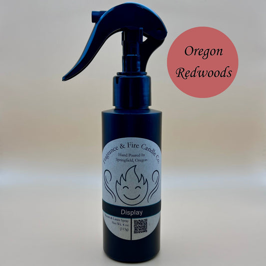 Black spray bottle from Fragrance & Fire Candle Co., Springfield, Oregon, with a black spray nozzle. The bottle has a white label with a stylized flame logo, the text "Fragrance & Fire Candle Co. Hand Poured In Springfield, Oregon," and "Display" scent name. The label also includes a QR code and the text "Room & Linen Spray Net Wt. 4 oz (113g)." A burnt red circle in the upper right corner reads "Oregon Redwoods."
