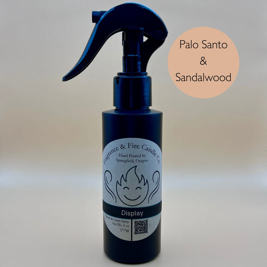 Black spray bottle from Fragrance & Fire Candle Co., Springfield, Oregon, with a black spray nozzle. The bottle has a white label with a stylized flame logo, the text "Fragrance & Fire Candle Co. Hand Poured In Springfield, Oregon," and "Display" scent name. The label also includes a QR code and the text "Room & Linen Spray Net Wt. 4 oz (113g)." A light brown circle in the upper right corner reads "Palo Santo & Sandalwood."