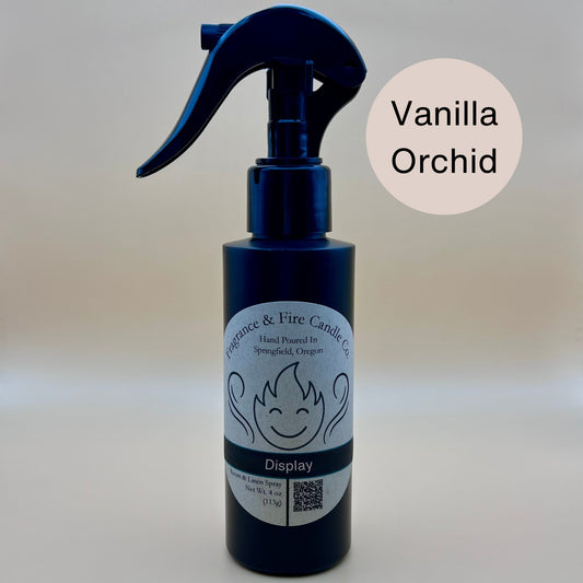 Black spray bottle from Fragrance & Fire Candle Co., Springfield, Oregon, with a black spray nozzle. The bottle has a white label with a stylized flame logo, the text "Fragrance & Fire Candle Co. Hand Poured In Springfield, Oregon," and "Display" scent name. The label also includes a QR code and the text "Room & Linen Spray Net Wt. 4 oz (113g)." A cream circle in the upper right corner reads "Vanilla Orchid."