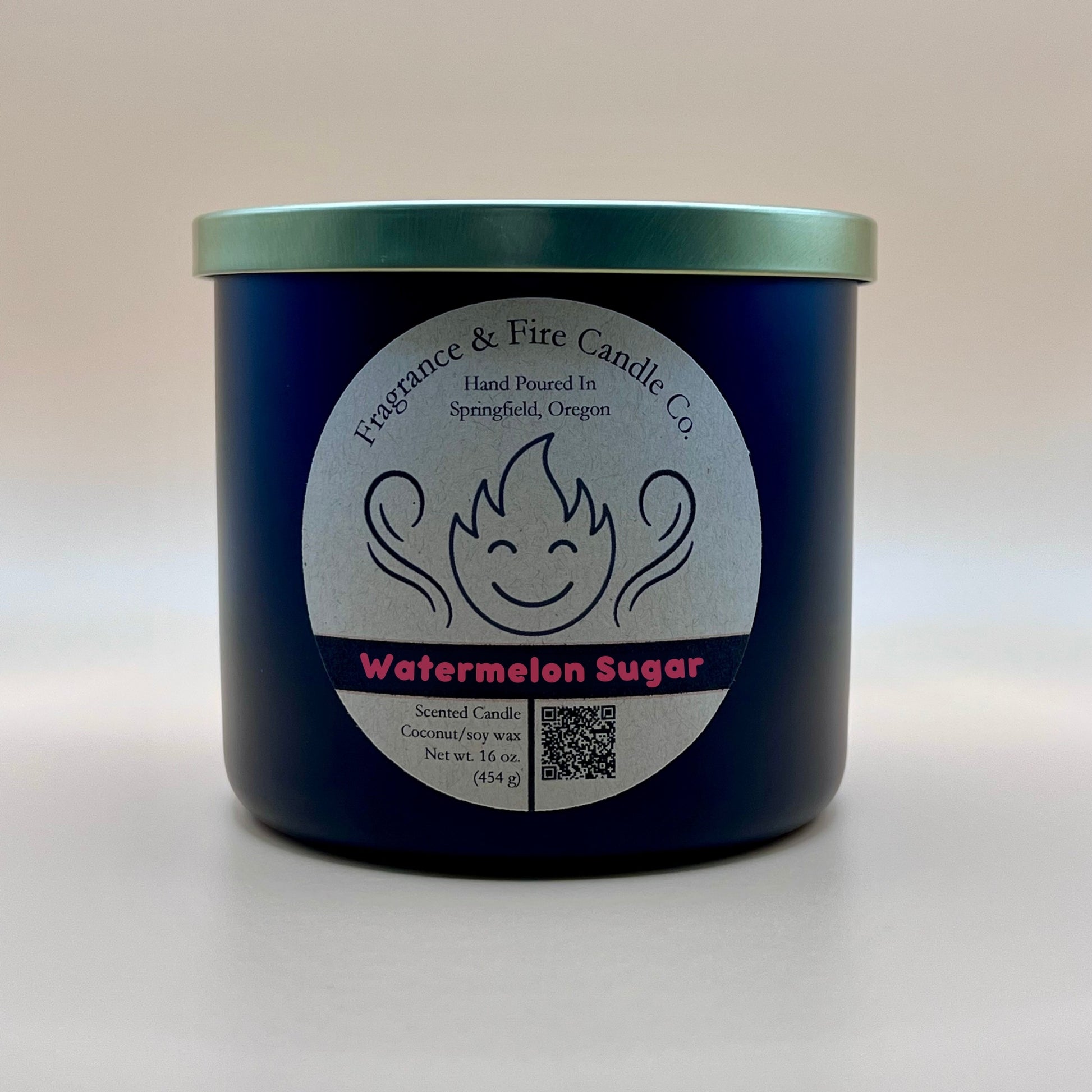 Black glass container candle from Fragrance & Fire Candle Co. with a gold lid. The label features a stylized flame with a smiling face, the text "Fragrance & Fire Candle Co. Hand Poured In Springfield, Oregon," and "Watermelon Sugar" scent name. Details include "Scented Candle Coconut/soy wax Net wt. 16 oz (454 g)" and a QR code.