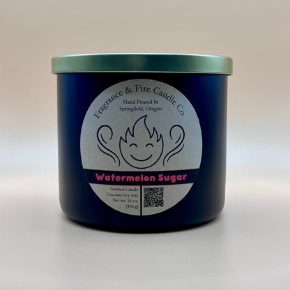 Black glass container candle from Fragrance & Fire Candle Co. with a gold lid. The label features a stylized flame with a smiling face, the text "Fragrance & Fire Candle Co. Hand Poured In Springfield, Oregon," and "Watermelon Sugar" scent name. Details include "Scented Candle Coconut/soy wax Net wt. 16 oz (454 g)" and a QR code.