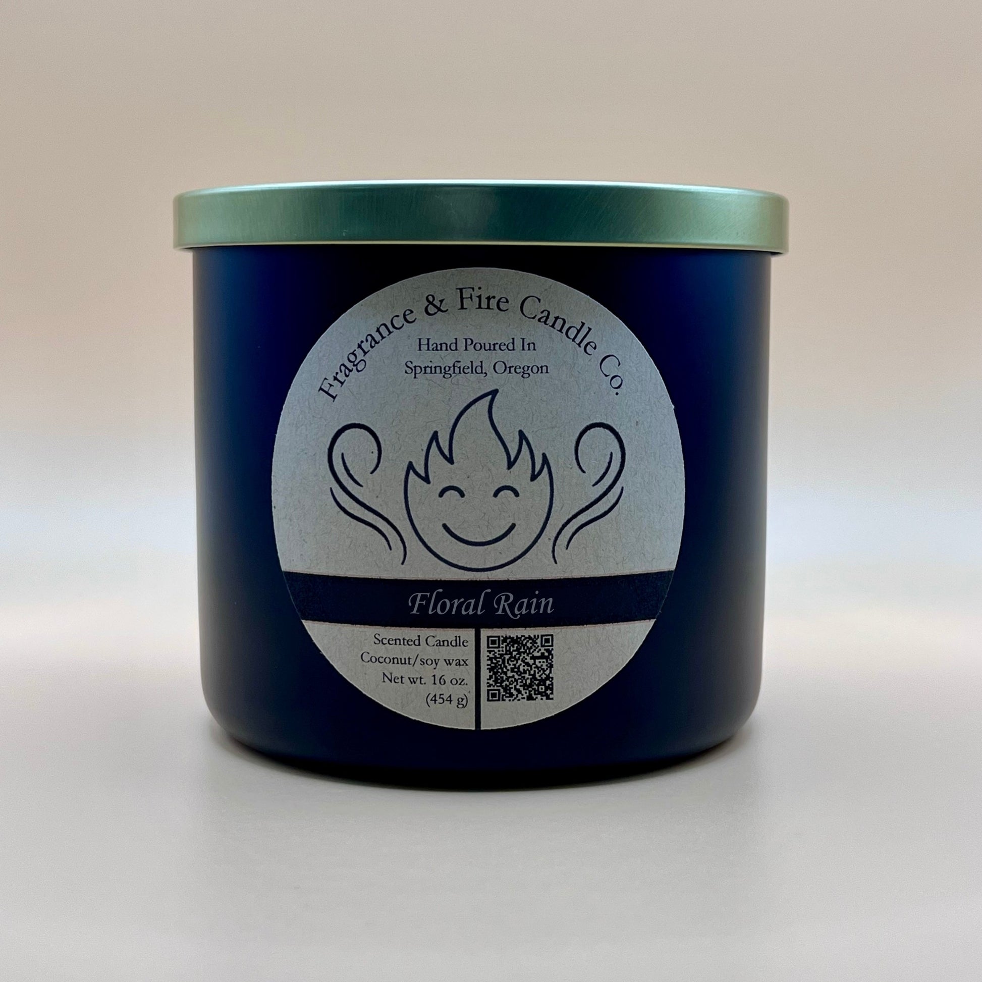 Black glass container candle from Fragrance & Fire Candle Co. with a gold lid. The label features a stylized flame with a smiling face, the text "Fragrance & Fire Candle Co. Hand Poured In Springfield, Oregon," and "Floral Rain" scent name. Details include "Scented Candle Coconut/soy wax Net wt. 16 oz (454 g)" and a QR code.