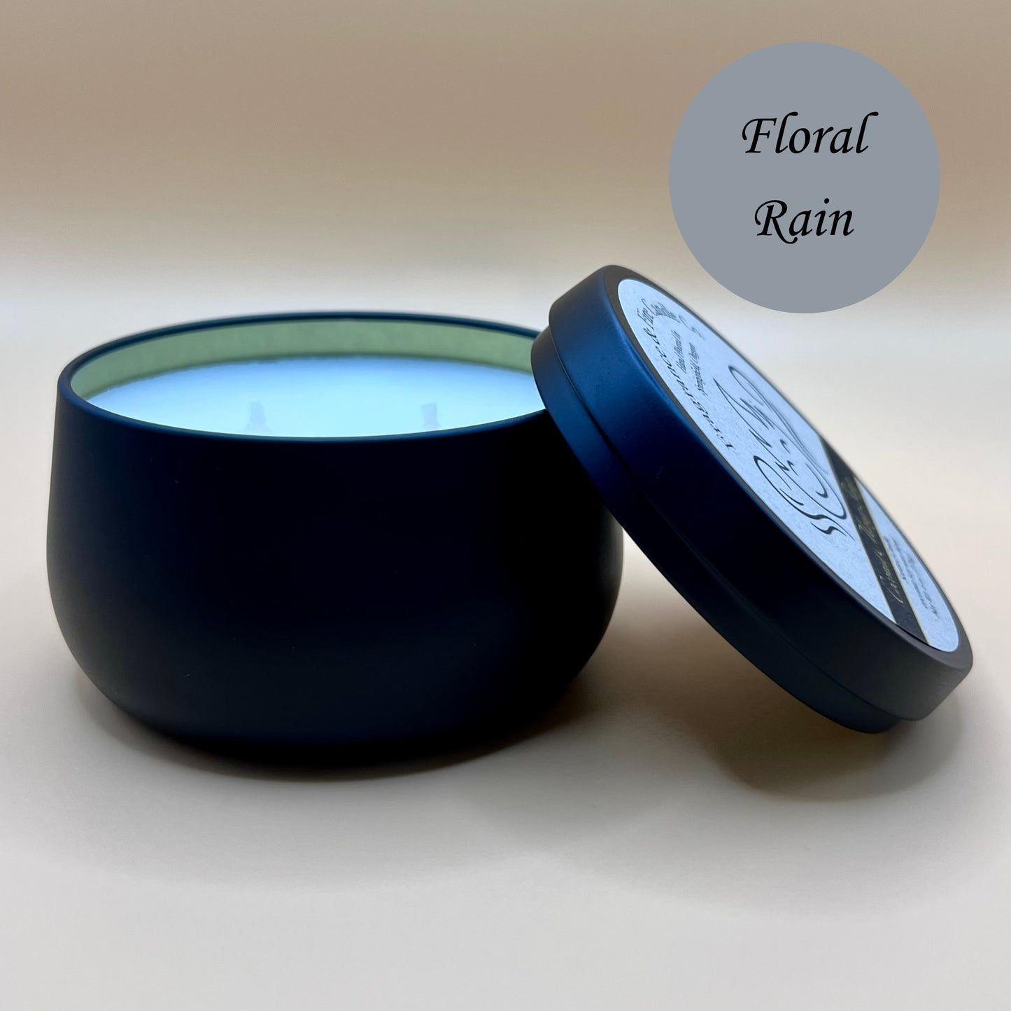 6oz black tin candle from Fragrance & Fire Candle Co., Springfield, Oregon, with an open lid resting beside it. The tin contains white wax. A teal circle in the upper right corner reads "Floral Rain." The open lid shows a partial label design.