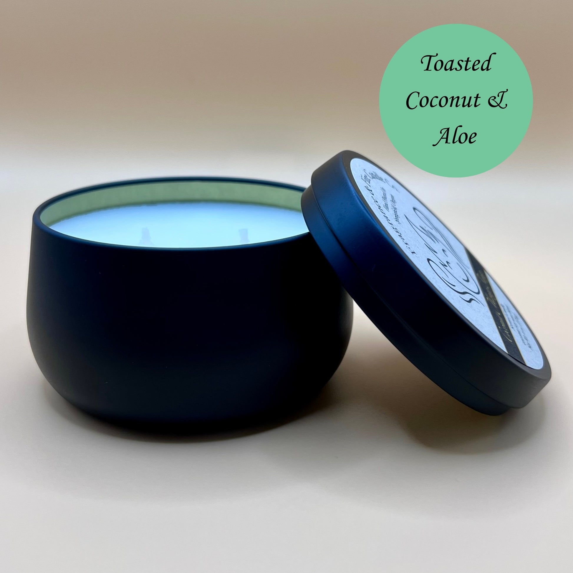6oz black tin candle from Fragrance & Fire Candle Co., Springfield, Oregon, with an open lid resting beside it. The tin contains white wax. A green circle in the upper right corner reads "Toasted Coconut & Aloe." The open lid shows a partial label design.