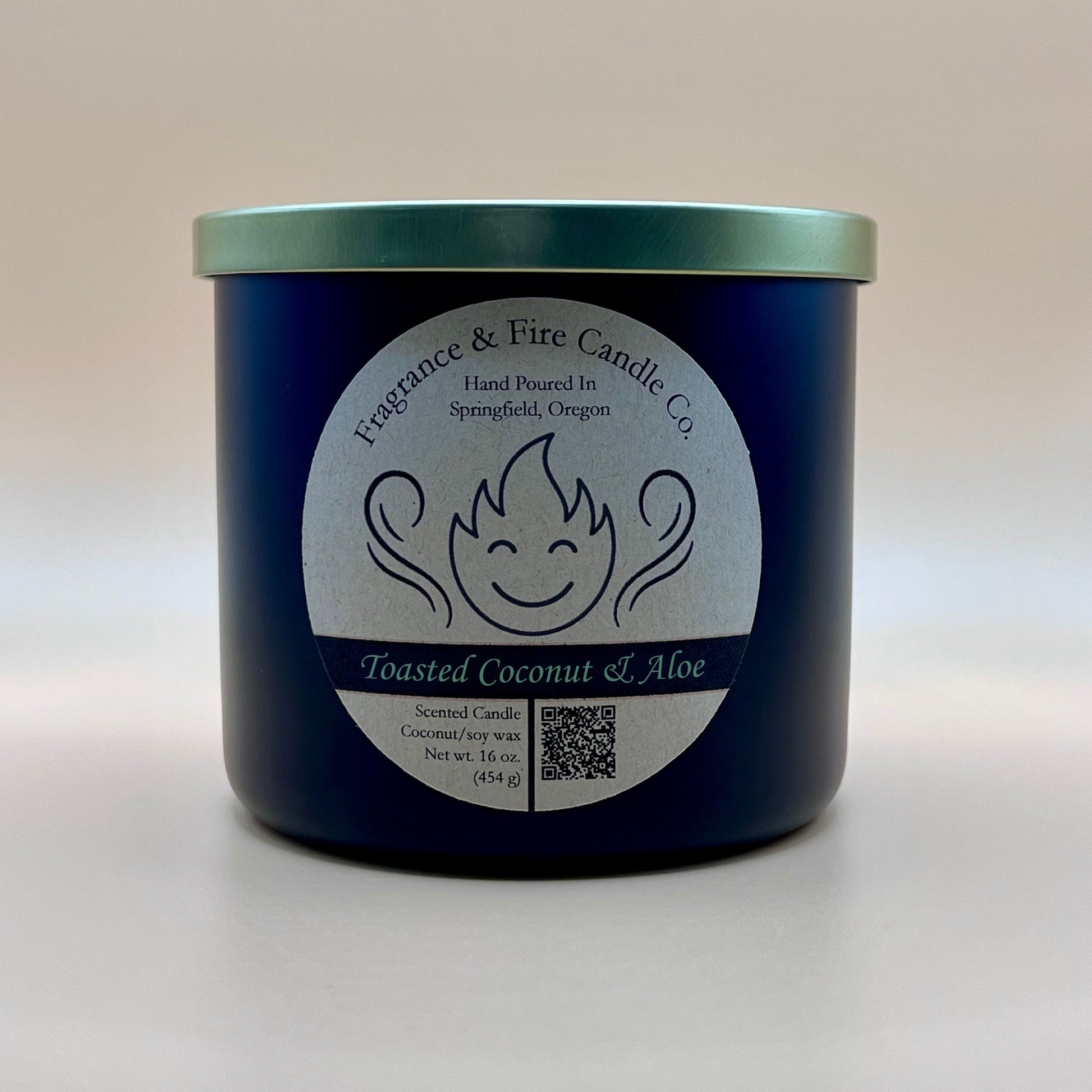 Black glass container candle from Fragrance & Fire Candle Co. with a gold lid. The label features a stylized flame with a smiling face, the text "Fragrance & Fire Candle Co. Hand Poured In Springfield, Oregon," and "Toasted Coconut & Aloe" scent name. Details include "Scented Candle Coconut/soy wax Net wt. 16 oz (454 g)" and a QR code.