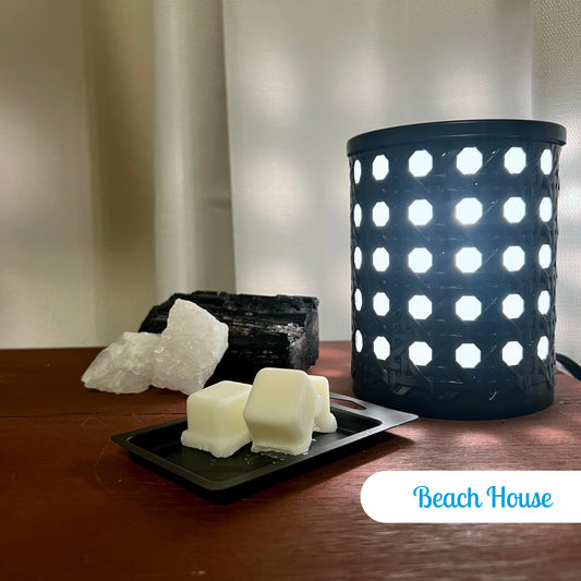 Beach House Wax Melts with three cubes in the lid next to a black, illuminated electric wax burner. Rocks are placed nearby, with a wall and curtain in the background.