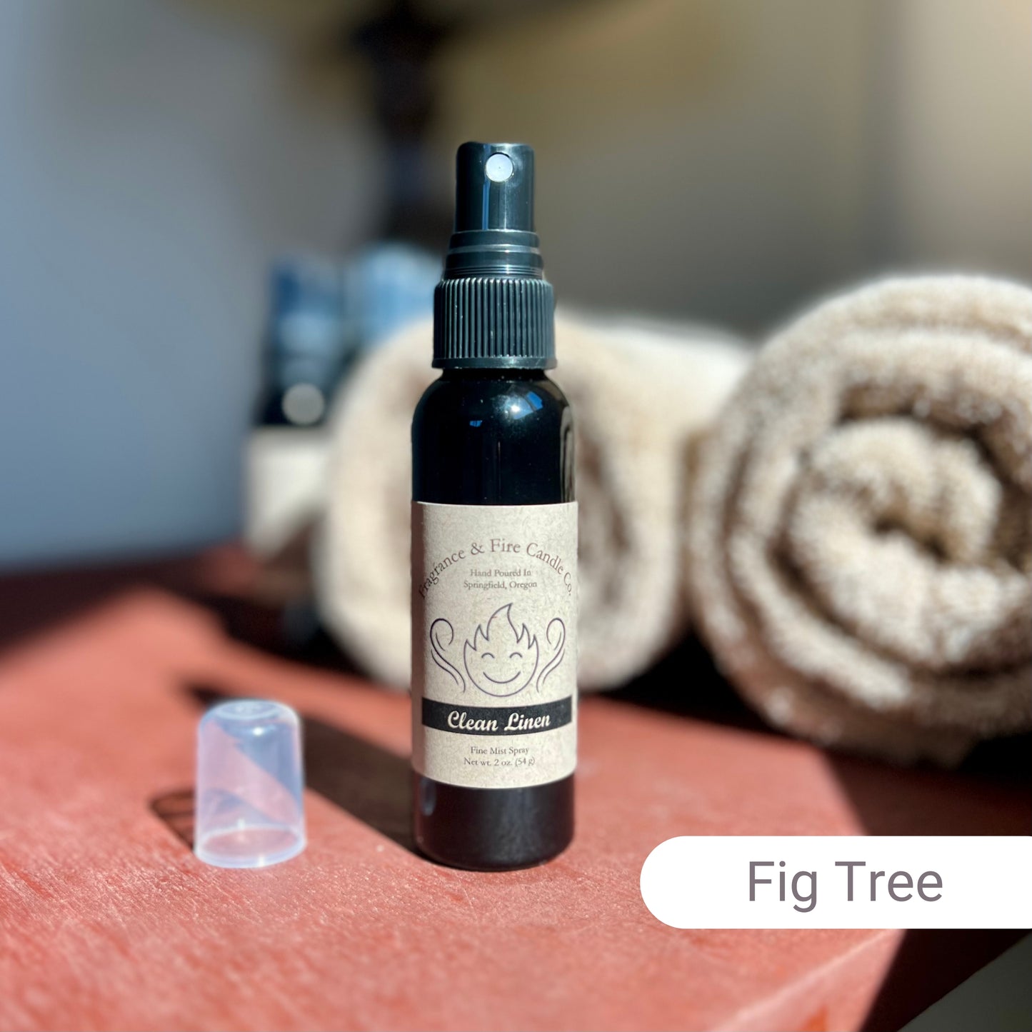 Fragrance & Fire Fig Tree Fragrance Mist in a sleek black bottle with a brown label, placed on a sunlit nightstand. The background features blurred brown towels and other sprays.