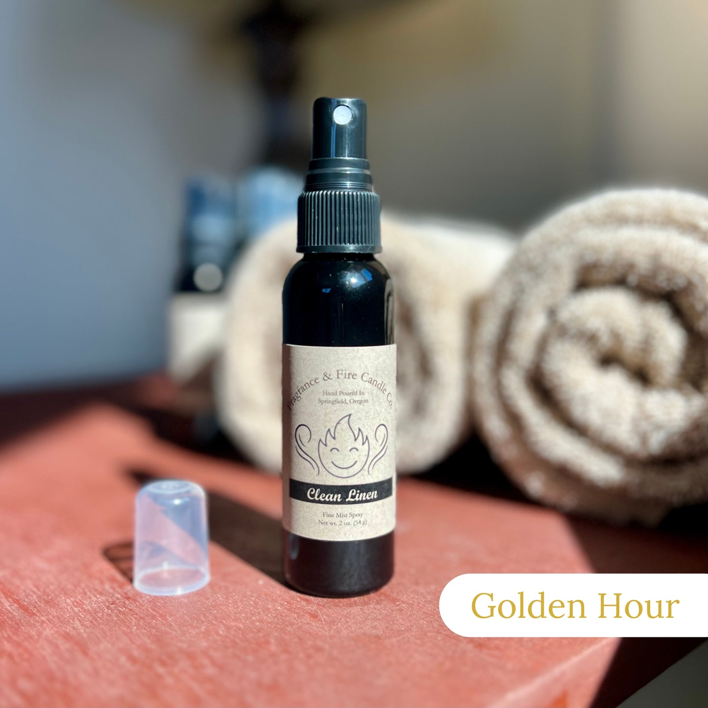 Fragrance & Fire Golden Hour Fragrance Mist in a sleek black bottle with a brown label, placed on a sunlit nightstand. The background features blurred brown towels and other sprays.