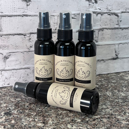 4 Fragrance & Fire Fragrance Mists in sleek black bottles. Brown label with company name and logo. 3 upright, 1 lying sideways in front. On granite surface with gray colored brick wall background.