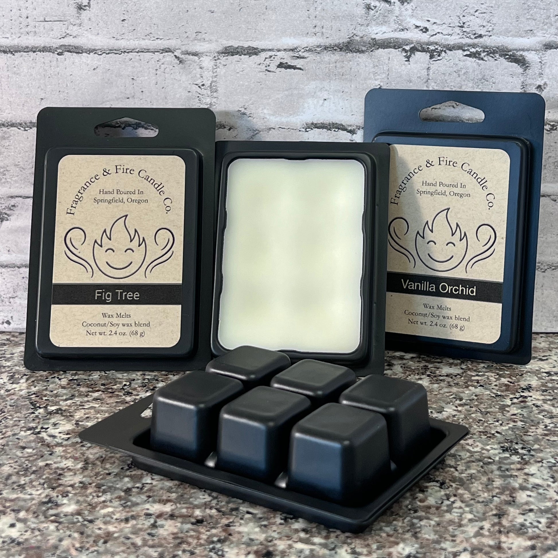 4 black packs Fragrance & Fire wax melts. Brown labels with company logo clearly visible, 1 no lid, 1 upside down. Sitting on granite countertop with grayish colored brick wall baskground.