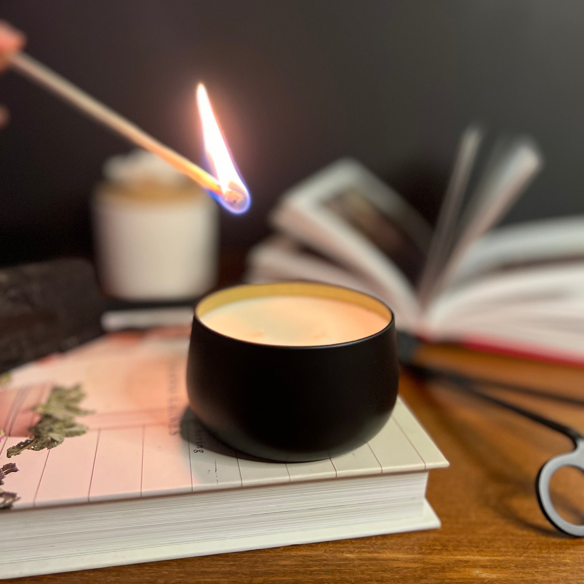 Fragrance & Fire candle on a book next to wick trimmers being lit by a match. Blurred book and diffuser in background. 