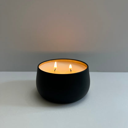 Black container candle, gold lining, 2 wicks lit, white background.