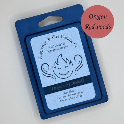 Black clamshell packaging for wax melts from Fragrance & Fire Candle Co., Springfield, Oregon. The packaging features a white label with a stylized flame logo, the text "Fragrance & Fire Candle Co. Hand Poured In Springfield, Oregon," and "Oregon Redwoods" scent name. The label also includes the text "Wax Melts Coconut/Soy wax blend Net wt. 2.5 oz. (70 g)." A burnt red circle in the upper right corner says scent name.