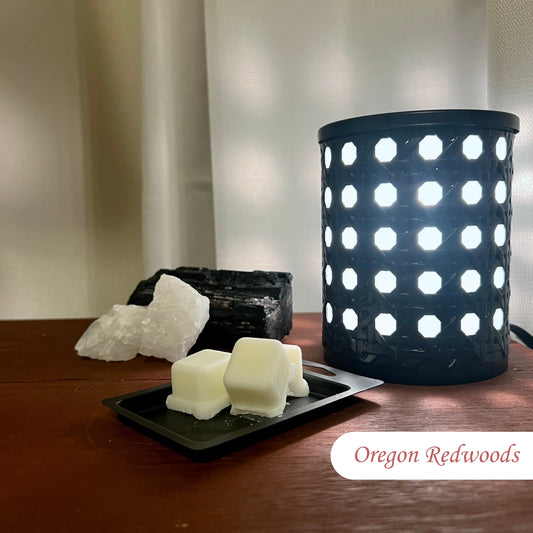 Oregon Redwoods Wax Melts with three cubes in the lid next to a black, illuminated electric wax burner. Rocks are placed nearby, with a wall and curtain in the background.