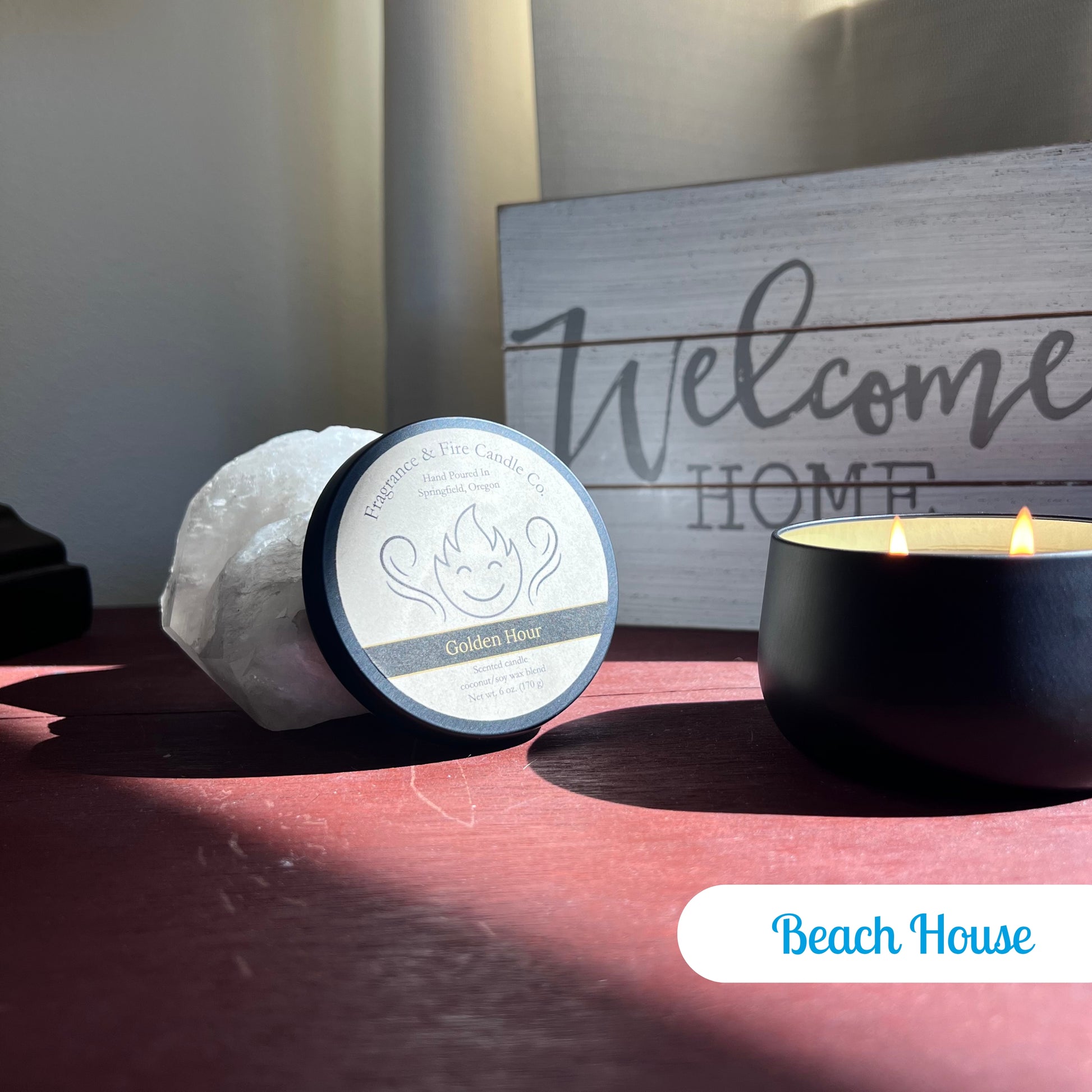 Lit Beach House Candle in a round black container on a bedside table with sunlight streaming through the window. The candle lid is propped against a crystal, and a 'Welcome Home' sign is visible in the background.