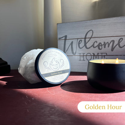 Lit Golden Hour Candle in a round black container on a bedside table with sunlight streaming through the window. The candle lid is propped against a crystal, and a 'Welcome Home' sign is visible in the background.