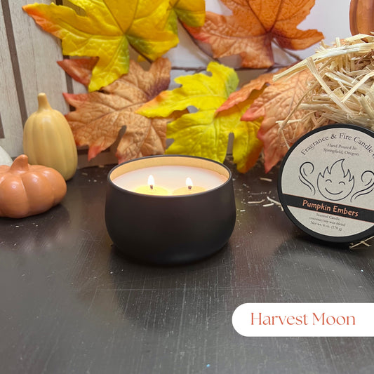 Lit Harvest Moon Candle in a round black container surrounded by fall decor, with the lid leaning against hay and the label clearly visible.