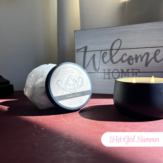 Lit Hot Girl Summer Candle in a round black container on a bedside table with sunlight streaming through the window. The candle lid is propped against a crystal, and a 'Welcome Home' sign is visible in the background.