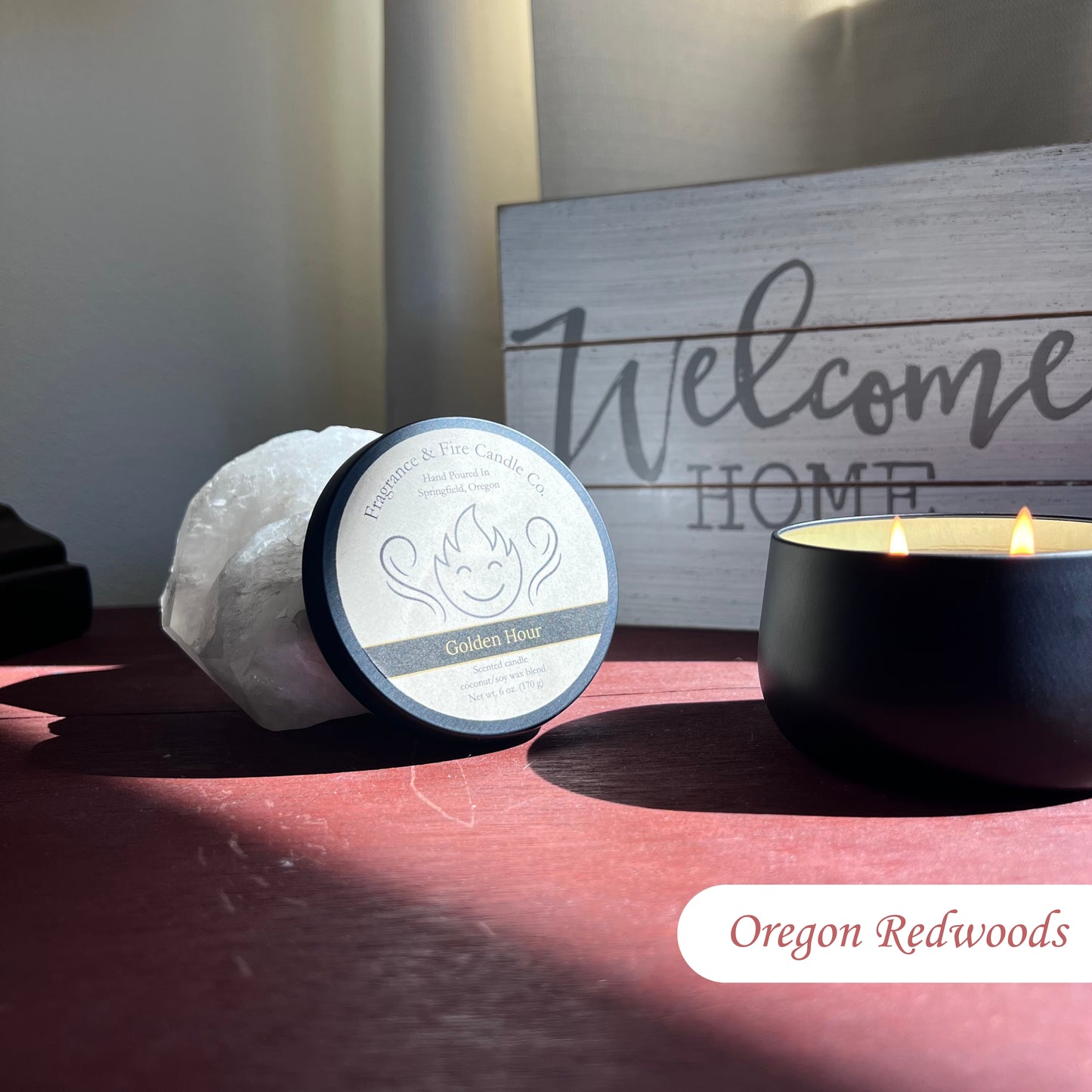 Lit Oregon Redwoods Candle in a round black container on a bedside table with sunlight streaming through the window. The candle lid is propped against a crystal, and a 'Welcome Home' sign is visible in the background.