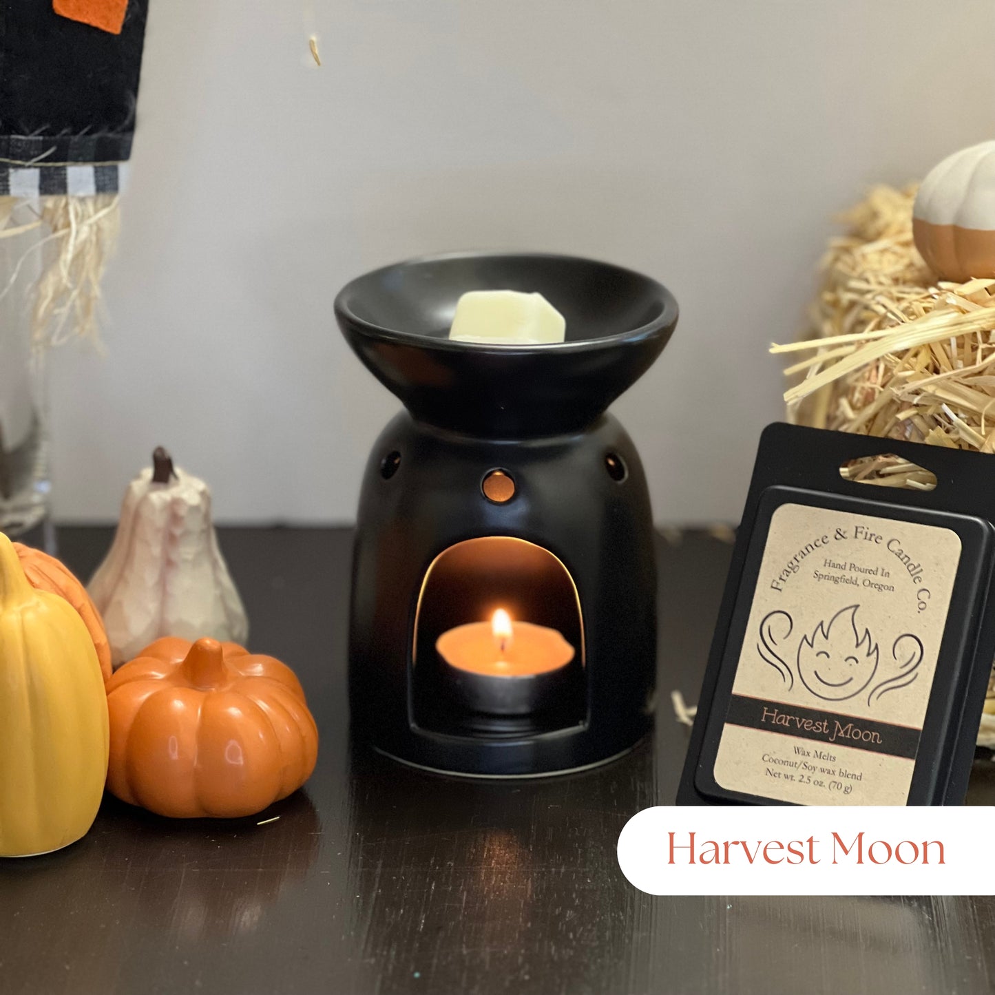 Harvest Moon Wax Melts with a partially melted cube in a tea light wax burner, lit with a tea light candle. Surrounded by fall decor including hay and gourds, with a pack of wax melts displayed upright by the melter.