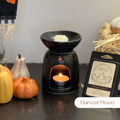 Harvest Moon Wax Melts with a partially melted cube in a tea light wax burner, lit with a tea light candle. Surrounded by fall decor including hay and gourds, with a pack of wax melts displayed upright by the melter.