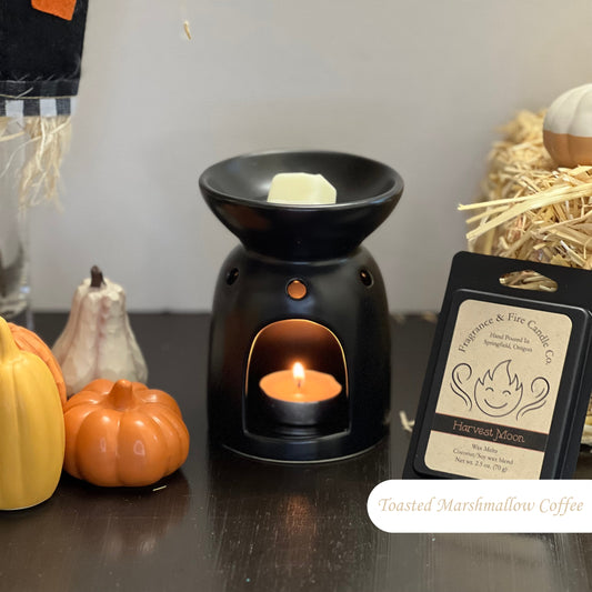 Toasted Marshmallow Coffee Wax Melts with a partially melted cube in a tea light wax burner, lit with a tea light candle. Surrounded by fall decor including hay and gourds, with a pack of wax melts displayed upright by the melter.